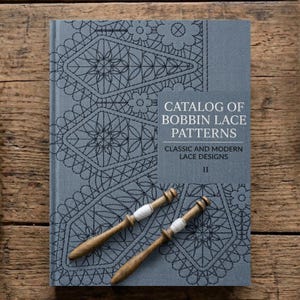 May include: A gray book titled "Catalog of Bobbin Lace Patterns" with classic and modern lace designs. The cover features intricate black lace patterns and two wooden bobbins with white thread. The book is on a wooden surface.