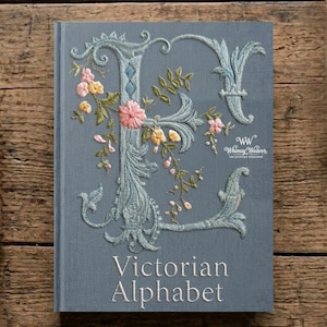 Pode incluir: Um livro azul-acinzentado intitulado "Victorian Alphabet" com uma letra "E" bordada adornada com flores rosa e amarelas e folhas verdes. A capa do livro apresenta o logotipo "Whimsy Weaver".
