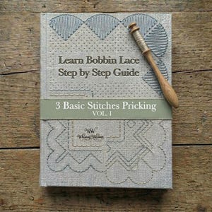 Peut inclure: Un livre à couverture rigide gris intitulé Learn Bobbin Lace Step by Step Guide avec une bande verte marquée 3 Basic Stitches Pricking VOL. I. Une bobine en bois avec du fil repose sur la couverture, ornée de motifs brodés et du logo Whimsy Weaver.