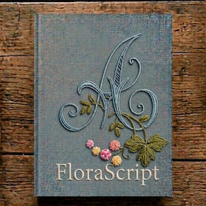 Op de afbeelding: Een grijs hardcover boek met een geborduurde grote cursieve letter A in lichtblauw garen. De letter is versierd met groene geborduurde bladeren en kleine roze en gele bloemaccenten. De tekst FloraScript is in het wit gedrukt.