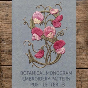 Può includere: Un tessuto azzurro con un ricamo floreale rosa e verde che forma la lettera "S". Il testo "BOTANICAL MONOGRAM EMBROIDERY PATTERN PDF - LETTER S" è sotto il disegno. Lo sfondo è una superficie di legno.