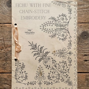 May include: A tan paper pattern booklet titled FICHU WITH FINE CHAIN-STITCH EMBROIDERY featuring black ink floral scrollwork designs and a logo for Whimsy Weaver. The booklet is bound on the left side with a light peach-colored satin ribbon.
