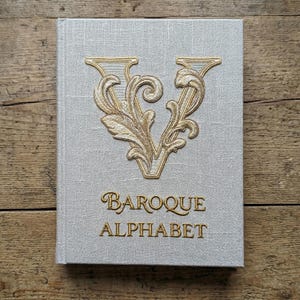 Könnte beinhalten: Ein Hardcover-Buch mit hellgrauem Leinenbezug. Das Cover zeigt einen goldgestickten Buchstaben "V" im Barockstil, darunter die gestickten Worte "Baroque Alphabet".