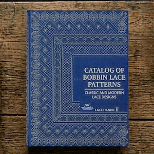 以下が含まれることがあります： 白いレースのデザインが施された、青い本「Catalog of Bobbin Lace Patterns」。表紙には幾何学模様と花柄が描かれています。本は木製の表面にあります。