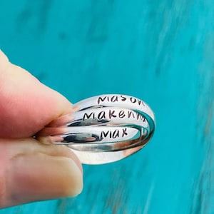 May include: Three silver rings stacked on top of each other. The rings are engraved with the names "Mason", "Makenna", and "Max".