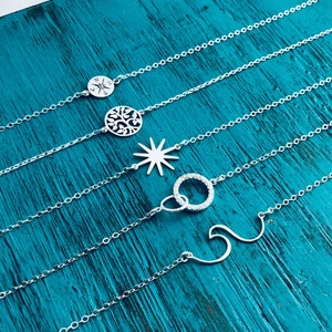May include: Five delicate silver necklaces with different pendant designs: a compass, a tree of life, a starburst, two interlocking circles with small clear stones, and a wave. The necklaces are displayed on a teal wooden surface.