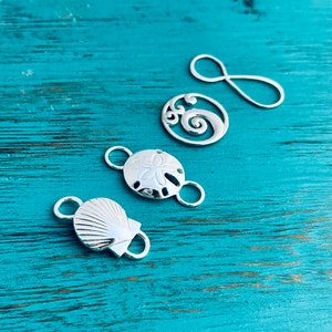 May include: Five silver-toned metal charms with beach-themed designs. The charms include a seashell, a starfish, a wave, and two infinity symbols.