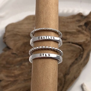 May include: Three silver rings stacked on a wooden surface. The top ring has a textured surface. The middle ring has a beaded design. The bottom ring has the name "RYAN" engraved on it. The ring above it has the name "CAITLIN" engraved on it.