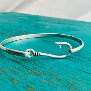 May include: A silver-colored bracelet with a hook and eye clasp. The bracelet is circular and made of a shiny metal. The hook clasp is shaped like a fish hook. The bracelet is displayed on a turquoise wooden surface.