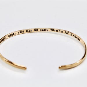 May include: Gold-colored open cuff bracelet. The bracelet has the engraved text: "BEAUTIFUL GIRL, YOU CAN DO HARD THINGS 🖤 BADASS". The jewelry is a simple, elegant design.