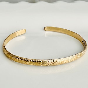 May include: A gold-toned bangle bracelet with an intricate floral design. The bracelet is engraved with the word "Aloha".