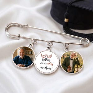 May include: A silver memorial pin with three round charms. Two charms feature photos of a man, and the center charm has the text "Walking with you today and Always!" with angel wings. A black graduation cap is in the background.
