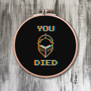 May include: A black embroidery hoop with the text "YOU DIED" in a pixelated font with rainbow colors. The text is surrounded by a pixelated image of a skull.