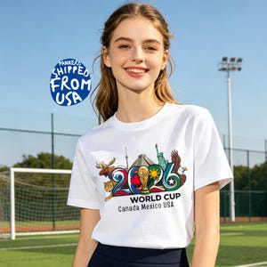 May include: White t-shirt featuring a colorful graphic design for the 2026 World Cup, with the text "Canada Mexico USA". The design includes a moose, a jaguar, the Eiffel Tower, the Statue of Liberty, and an eagle.