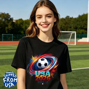 May include: Black t-shirt featuring a soccer ball design with red, blue, and yellow accents. The shirt has the text "USA 2026 World Cup" in blue and red. The shirt is worn by a person.