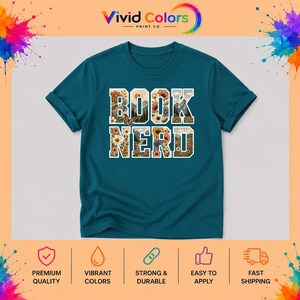 May include: A teal short-sleeved t-shirt featuring the text "BOOK NERD" in large block letters. The letters are filled with a graphic design of mountains, flowers, and a butterfly. The shirt has a crew neckline and a standard fit.