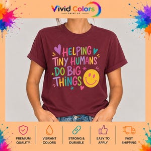 May include: A maroon short-sleeved t-shirt featuring the colorful text HELPING TINY HUMANS DO BIG THINGS in various fonts and colors, accompanied by small heart and star graphics and a large yellow smiley face icon on the front.