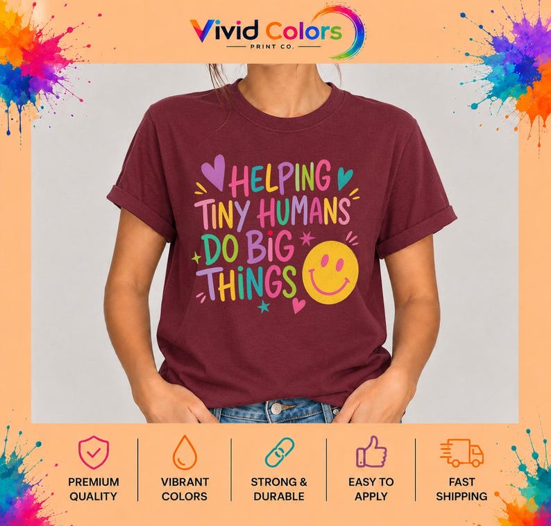May include: A maroon short-sleeved t-shirt featuring the colorful text HELPING TINY HUMANS DO BIG THINGS in various fonts and colors, accompanied by small heart and star graphics and a large yellow smiley face icon on the front.