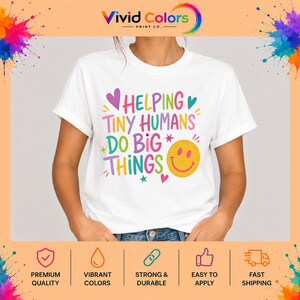 May include: A white short-sleeved t-shirt featuring the colorful text "HELPING TINY HUMANS DO BIG THINGS" in various fonts and shades of pink, yellow, teal, and purple, accompanied by a yellow smiley face icon and small heart and star accents.