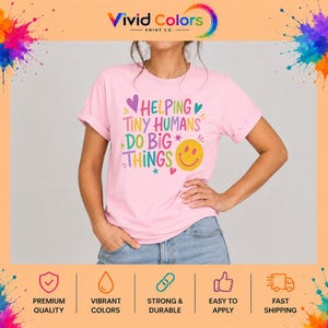 May include: A light pink short-sleeve t-shirt featuring the multi-colored text HELPING TINY HUMANS DO BIG THINGS. The design includes a yellow smiley face, small purple and teal hearts, and star accents printed on the front of the garment.