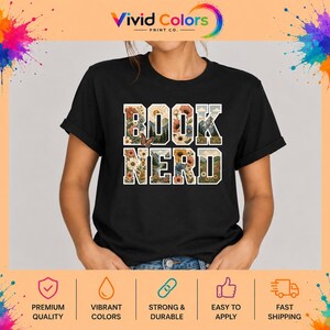 May include: A black short-sleeved t-shirt featuring the text BOOK NERD in large block letters. The letters are filled with a colorful graphic design of various flowers, green mountains, a sun, and a small butterfly on the letter O.