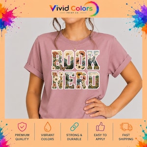 May include: A mauve short-sleeved t-shirt featuring the text BOOK NERD in large block letters. The letters are filled with a graphic design of colorful flowers, green trees, mountains, a sun, and a small butterfly perched on the letter K.