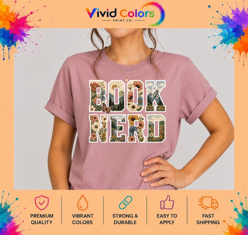 May include: A mauve short-sleeved t-shirt featuring the text BOOK NERD in large block letters. The letters are filled with a graphic design of colorful flowers, green trees, mountains, a sun, and a small butterfly perched on the letter K.