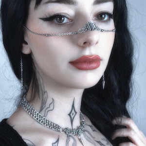 May include: A silver chainmail nose piece, necklace, and earrings set. The nose piece connects to the necklace with a delicate chain. The necklace features a spike pendant. The earrings are also spike shaped.