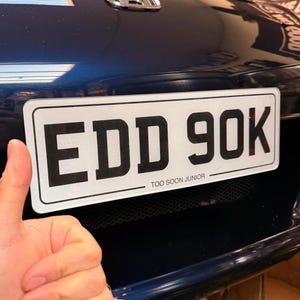 Puede incluir: Primer plano de la parte delantera de un coche azul oscuro con una matrícula blanca. La placa muestra las letras y números negros "EDD 90K" y el texto "TOO SOON JUNIOR" debajo.