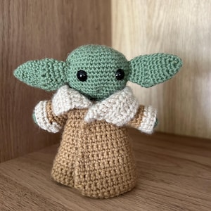 Könnte beinhalten: Handgemachte Häkelpuppe einer Figur mit großen Ohren, schwarzen Augen und einem beigefarbenen Gewand. Die Puppe ist grün, beige und cremefarben. Die Puppe ist etwa 15 cm groß.