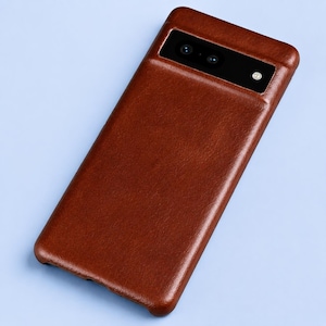 May include: A brown leather smartphone case with a smooth, textured finish. The case features a rectangular cutout at the top for the camera module, which contains two circular lenses and a small circular flash component on the right side.