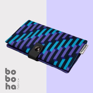 Pode incluir: Uma carteira retangular de tecido com um padrão geométrico de barras repetitivas em azul escuro, roxo e ciano. A carteira é fixada com uma alça preta e um botão de pressão de metal prateado. Uma pequena etiqueta de tecido branca contém o texto boboha.