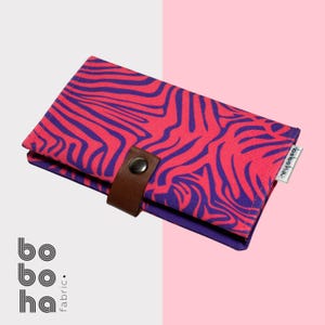 Puede incluir: Una cartera de tela rectangular con un vibrante patrón ondulado de color rosa y morado similar al de una cebra. La cartera se asegura con una correa de piel sintética marrón y un botón de presión metálico plateado. Incluye una etiqueta boboha.