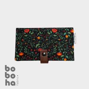 Op de afbeelding: Een rechthoekige stoffen portemonnee met een zwarte achtergrond en een dicht patroon van rode bloemen, oranje knoppen en groen blad. De portemonnee is voorzien van een bruin leren lipje met een metalen drukknoop en een klein wit label.