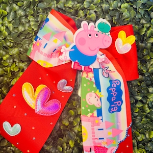 Puede incluir: Un lazo rojo grande para el cabello con Peppa Pig y una rana. El lazo tiene un diseño de castillo colorido y detalles de corazón. El texto "Peppa Pig" es visible en el lazo.