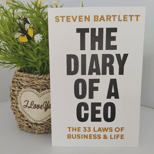 Peut inclure: Un livre intitul&eacute; "The Diary of a CEO" de Steven Bartlett, avec une couverture blanche et un grand texte noir. Le sous-titre est "The 33 Laws of Business & Life". Un plante d&eacute;corative dans un panier en osier est en arri&egrave;re-plan.