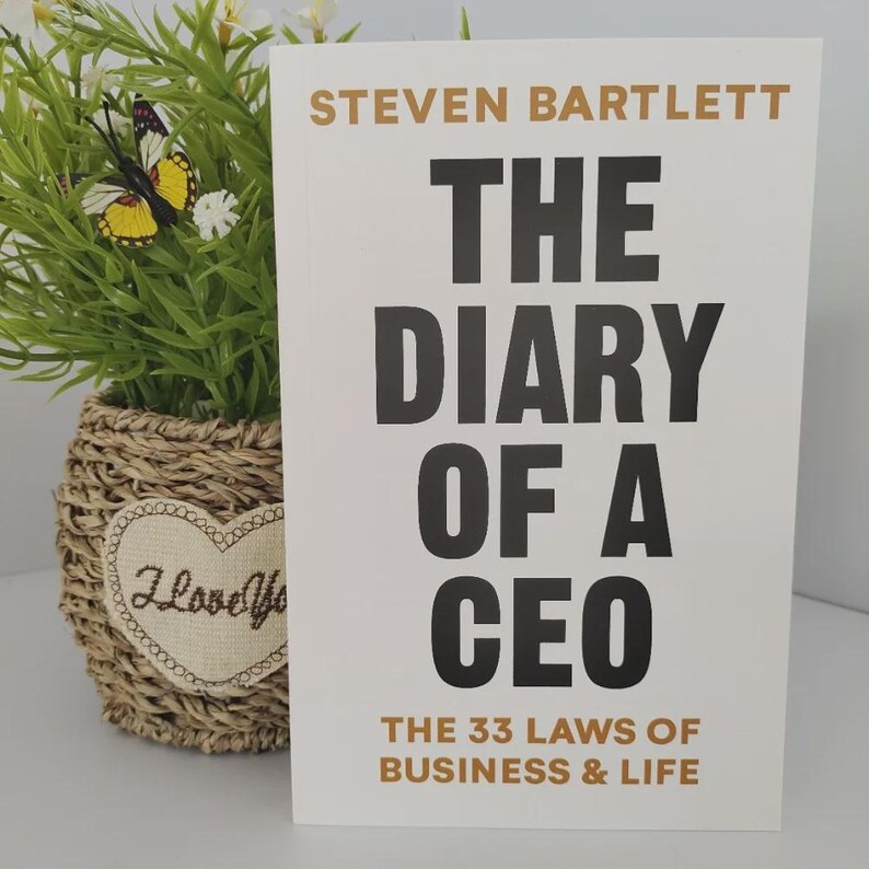 Peut inclure: Un livre intitul&eacute; "The Diary of a CEO" de Steven Bartlett, avec une couverture blanche et un grand texte noir. Le sous-titre est "The 33 Laws of Business & Life". Un plante d&eacute;corative dans un panier en osier est en arri&egrave;re-plan.