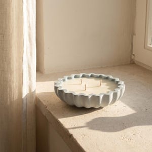 Peut inclure: Une bougie ronde dans un récipient bleu clair avec un bord festonné et ondulé. Le contenant est rempli de cire lisse blanc cassé et comporte trois mèches verticales positionnées au centre. Le design est simple, circulaire et mat.