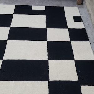 Pode incluir: Um tapete retangular com um padrão de xadrez de alto contraste, composto por quadrados alternados em preto e branco sujo. O tapete possui uma superfície texturizada e tufada com bordas retas e limpas, criando um design geométrico marcante.