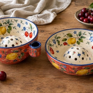 May include: Two ceramic berry bowls with a red exterior and cream interior, featuring hand-painted yellow lemons, red cherries, and blue flowers. Each bowl has a central perforated dome for rinsing and a small side handle for easy gripping.