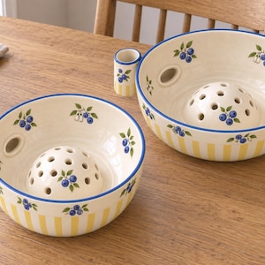 May include: Two round ceramic bowls with a cream-coloured base, featuring a yellow vertical striped pattern on the exterior and a hand-painted blueberry motif. Each bowl includes a central dome with drainage holes and a small side cylinder attachment.