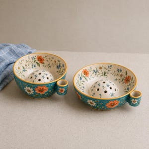 Puede incluir: Dos cuencos de cerámica con exterior azul turquesa e interior crema, decorados con motivos florales naranjas y blancos. Cada cuenco incluye una cúpula central elevada con pequeñas perforaciones circulares y una pequeña asa lateral.