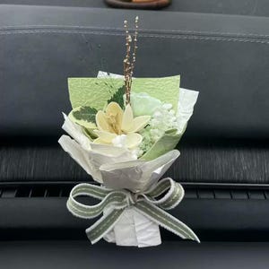 May include: A small, decorative bouquet featuring a cream-colored orchid and white flowers, wrapped in white paper and tied with a green and white striped ribbon. The bouquet includes green leaves and stems, creating a natural aesthetic.