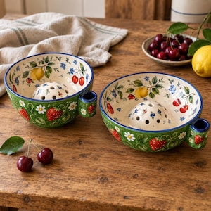 Peut inclure: Deux bols à baies en céramique avec un extérieur vert et un intérieur crème, décorés de fraises, cerises, citrons et marguerites blanches peints. Chaque bol possède un dôme central perforé pour le rinçage et une petite poignée latérale.
