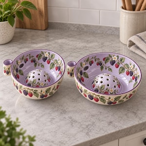 May include: Two round ceramic berry bowls with a light purple interior and a white exterior. Both bowls feature a central raised dome with drainage holes, a small side handle, and a decorative pattern of green leaves, white flowers, and red and black berries.