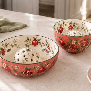 May include: Two ceramic bowls with a red exterior and cream interior, featuring a pomegranate and floral pattern. Each bowl has a central perforated dome and a small side handle. The design includes red pomegranates and green leaves.