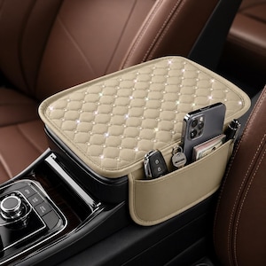 Puede incluir: Organizador de consola de coche beige con una parte superior acolchada y bolsillos laterales. El organizador contiene un teléfono inteligente, llaves, dinero en efectivo y un bolígrafo. El interior del coche es marrón.