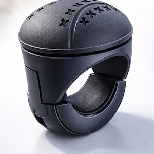 May include: A matte black, circular device with a perforated top and a ring-like base. The top features a series of cross-shaped cutouts. The device appears to be made of a durable material and is designed for a specific purpose.