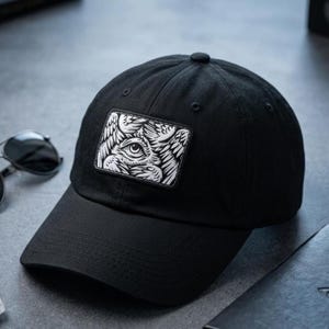 May include: A black baseball cap featuring a rectangular patch on the front. The patch displays a detailed black and white graphic illustration of a single eye surrounded by multiple feathered wings, creating a layered and intricate visual design.