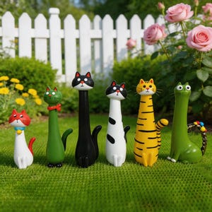 Puede incluir: Una colección de seis figuras de gatos caprichosos con cuellos alargados, cada uno pintado con una combinación de colores única. Los gatos están colocados sobre un césped, con una valla blanca y rosales al fondo. Las figuras son de cerámica.